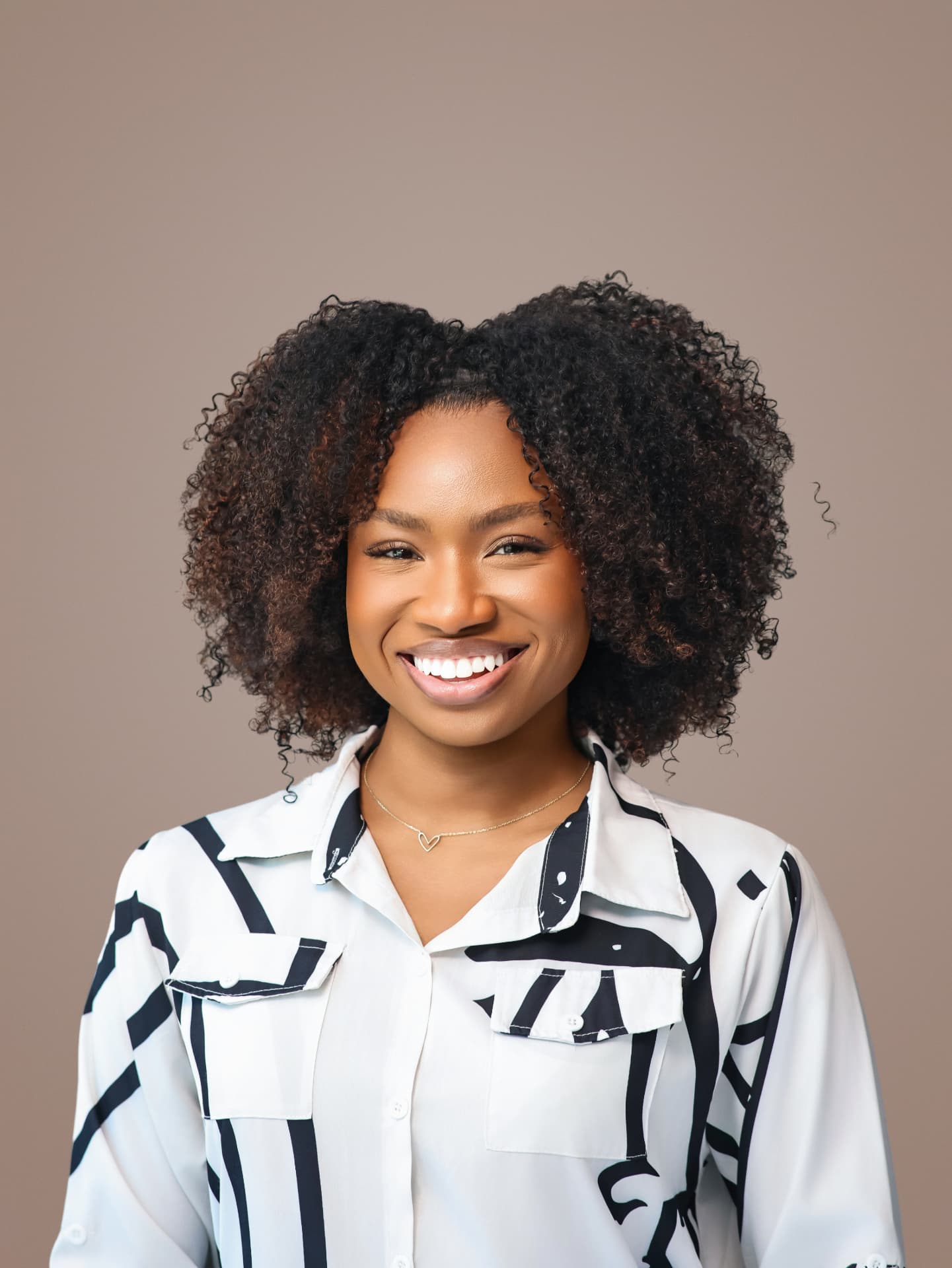 Smiling woman with curly hair in a black and white print shirt on a taupe backdrop