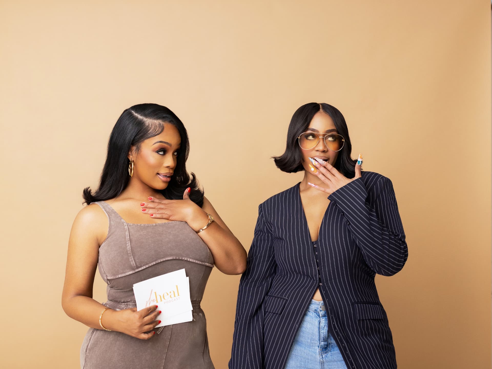 Two hosts laughing on a tan backdrop, one holding a cue card