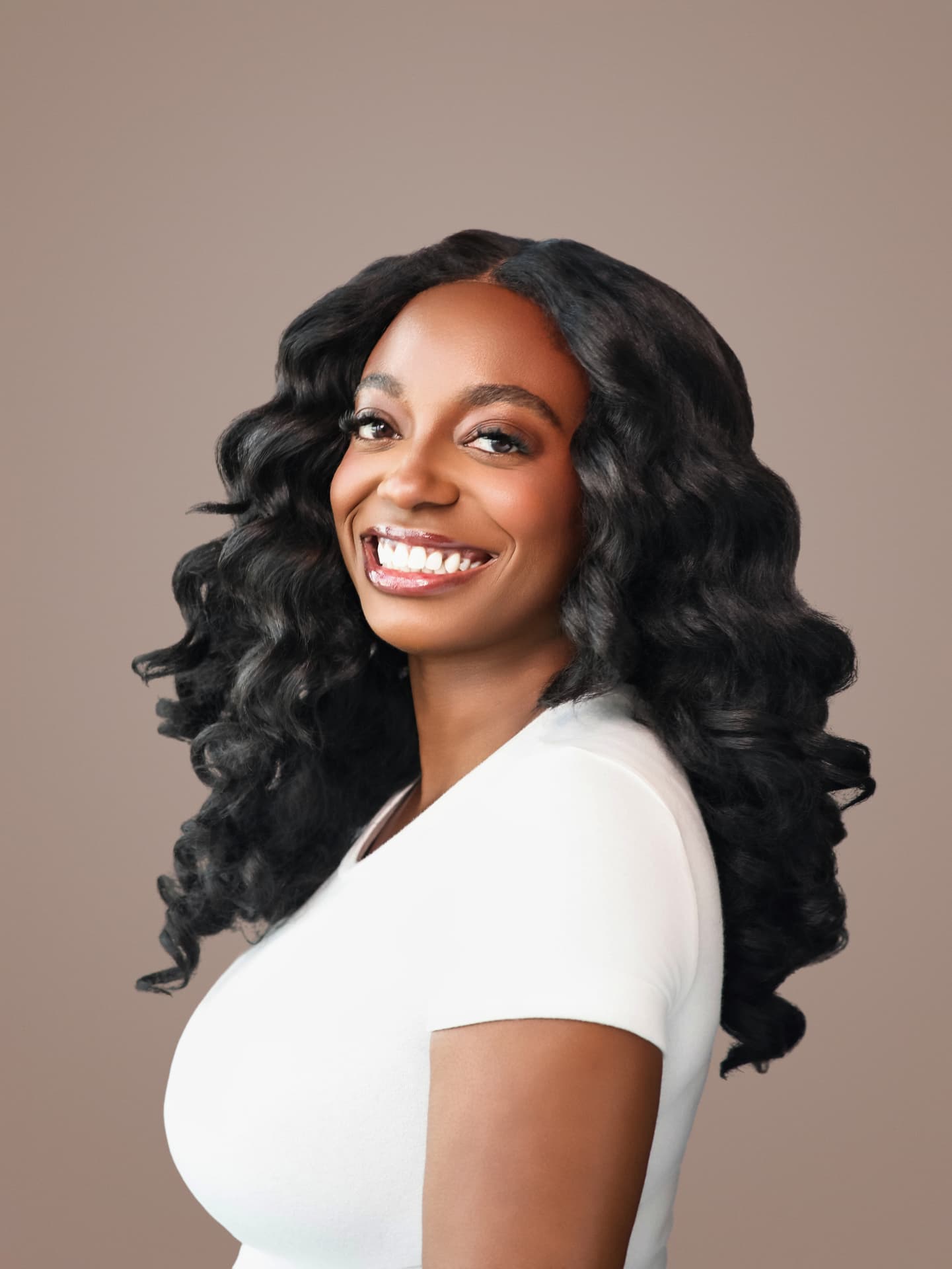 Smiling woman with long curls in a white tee on a taupe backdrop
