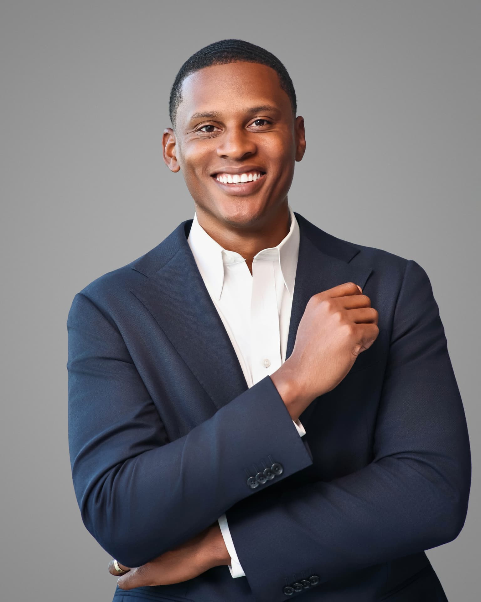 Smiling man in a navy blazer and white shirt with arms folded on a gray backdrop