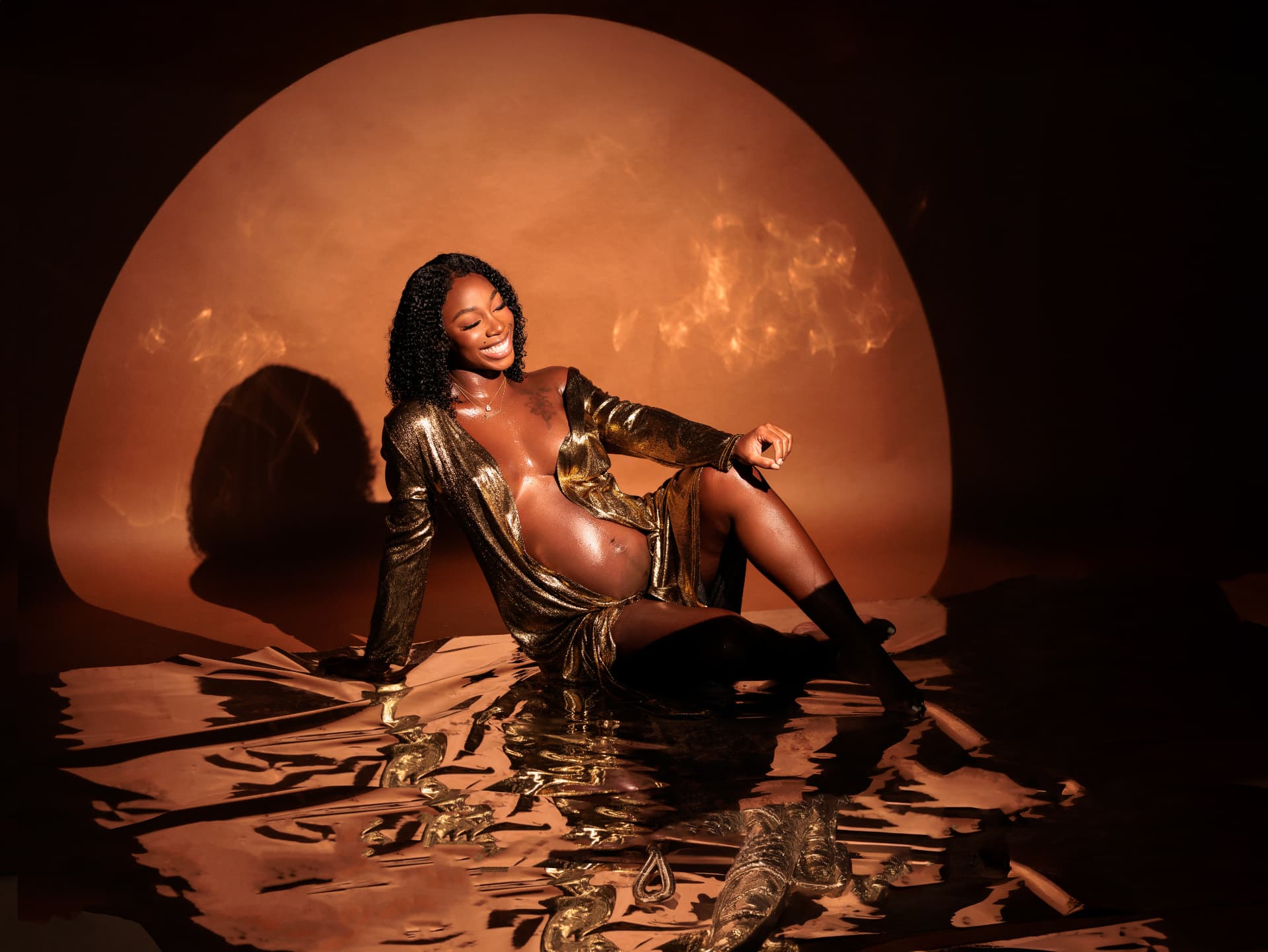 Smiling expectant mother in a gold metallic wrap seated on reflective gold under a warm spotlight