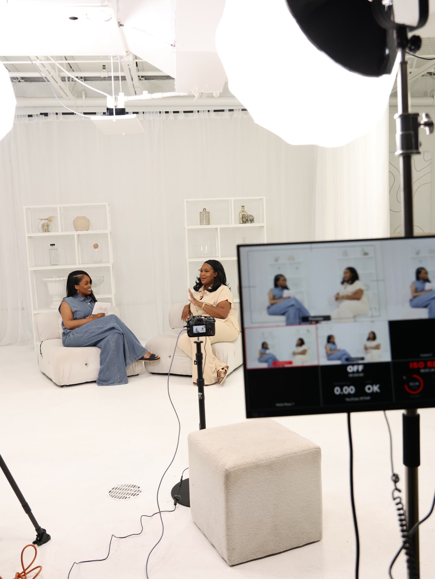 Behind the scenes of a podcast recording, two hosts on cream seating with lights and a monitor